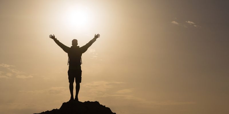 Man with arms outstretched celebrating or praying in beautiful inspiring sunrise with mountains and sea. Man hiking or climbing with hands up enjoy inspirational landscape on rocky top on Crete.